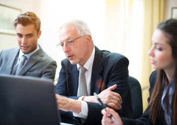 Drei Personen in Businesskleidung sitzen vor einem Laptop, der Mittlere erklärt etwas
